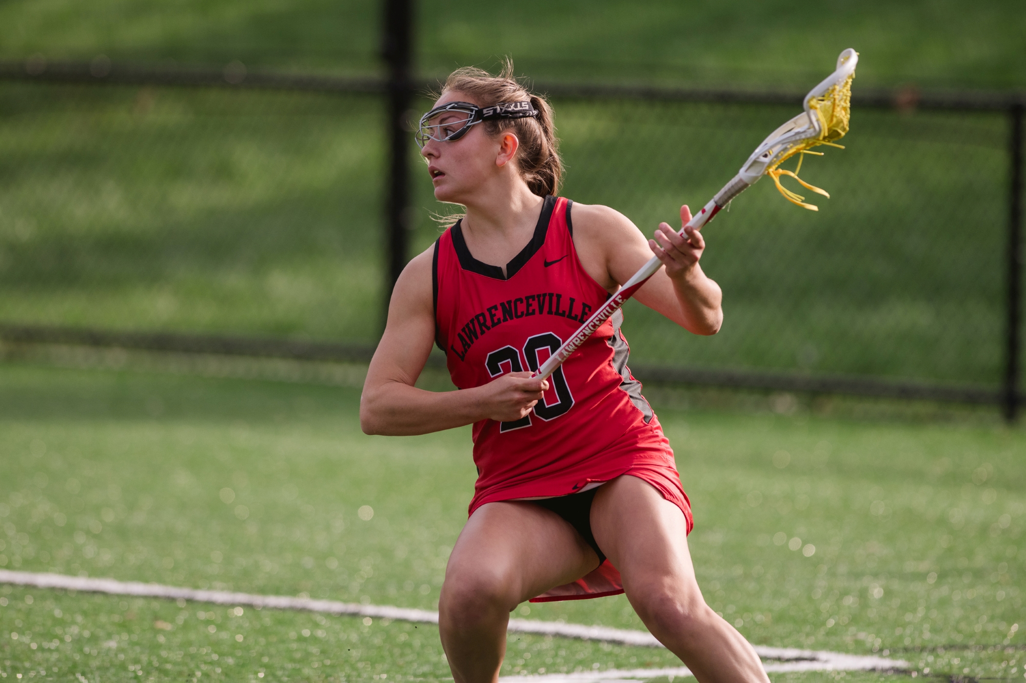 A girl in a red Lawrenceville lacrosse uniform eyes the play while carrying her stick, with the basket to the left.