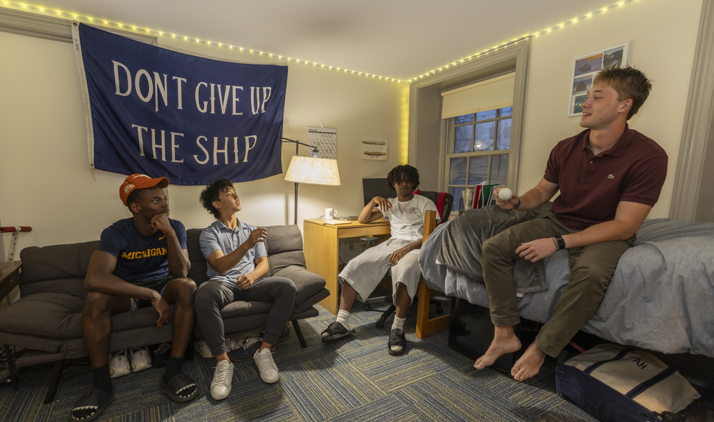 An older student sits on the top of a bunk bed in a dorm room, speaking, while three younger students pay attention to him.