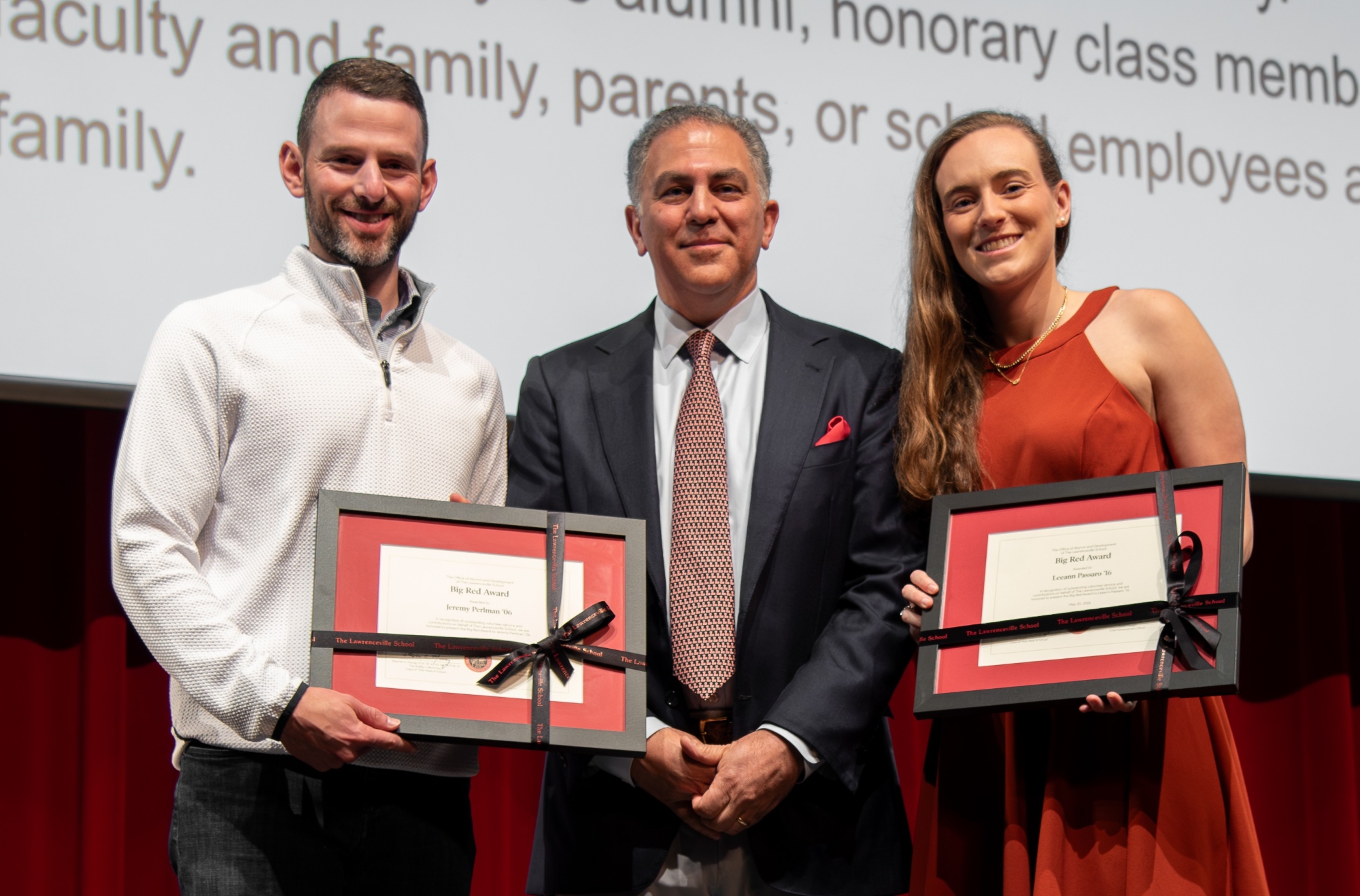 A man in a suit is flanked by a man in a white button-down shirt and a woman in a red dress. The people on the ends are holding framed certificates wrapped in black ribbons.