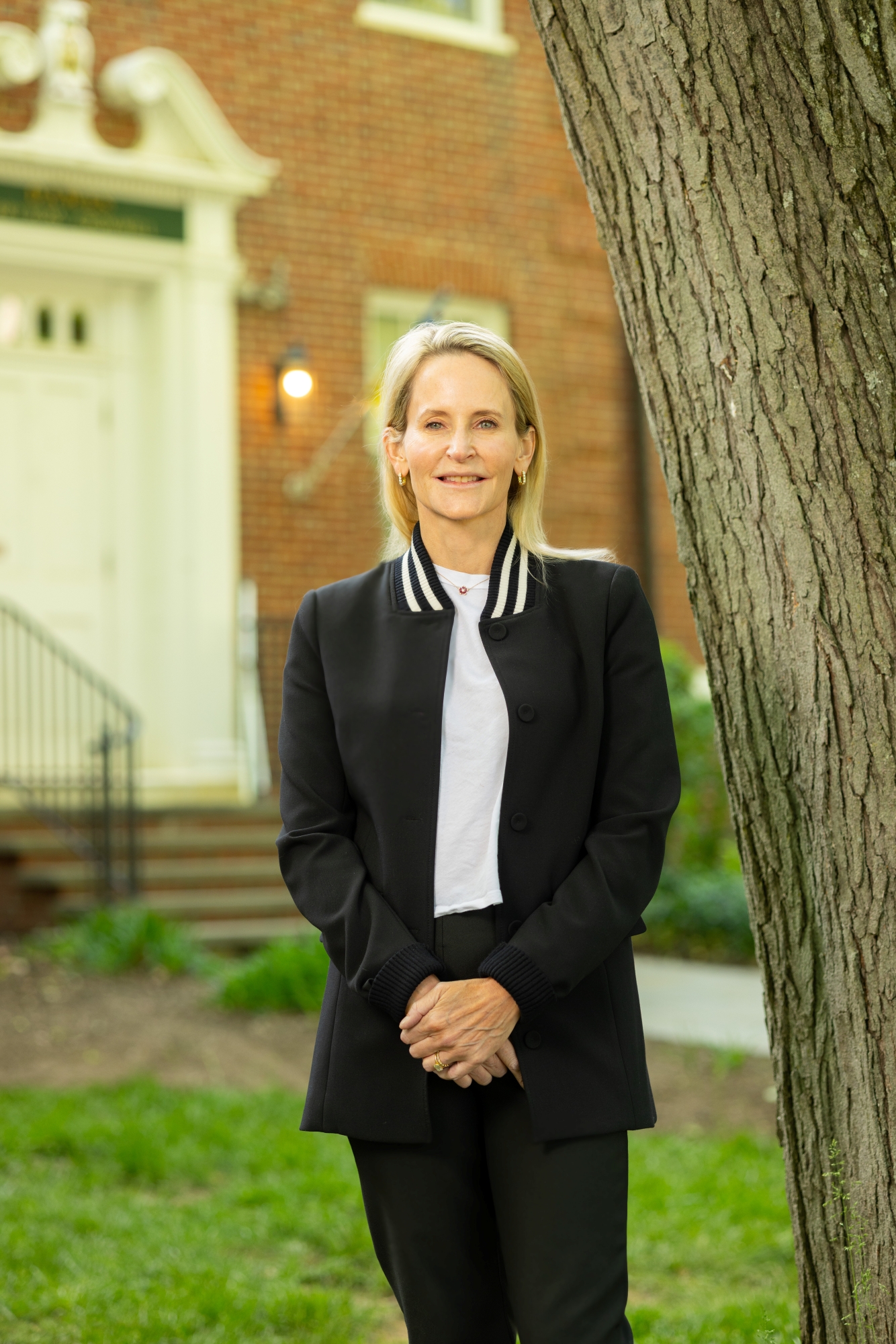 Celeste Mellet stands, smiling, with her hands clasped in front of her. She is wearing a black suit and a white shirt. She has shoulder-length blonde hair and a fair complexion. Behind her is a brick academic building.