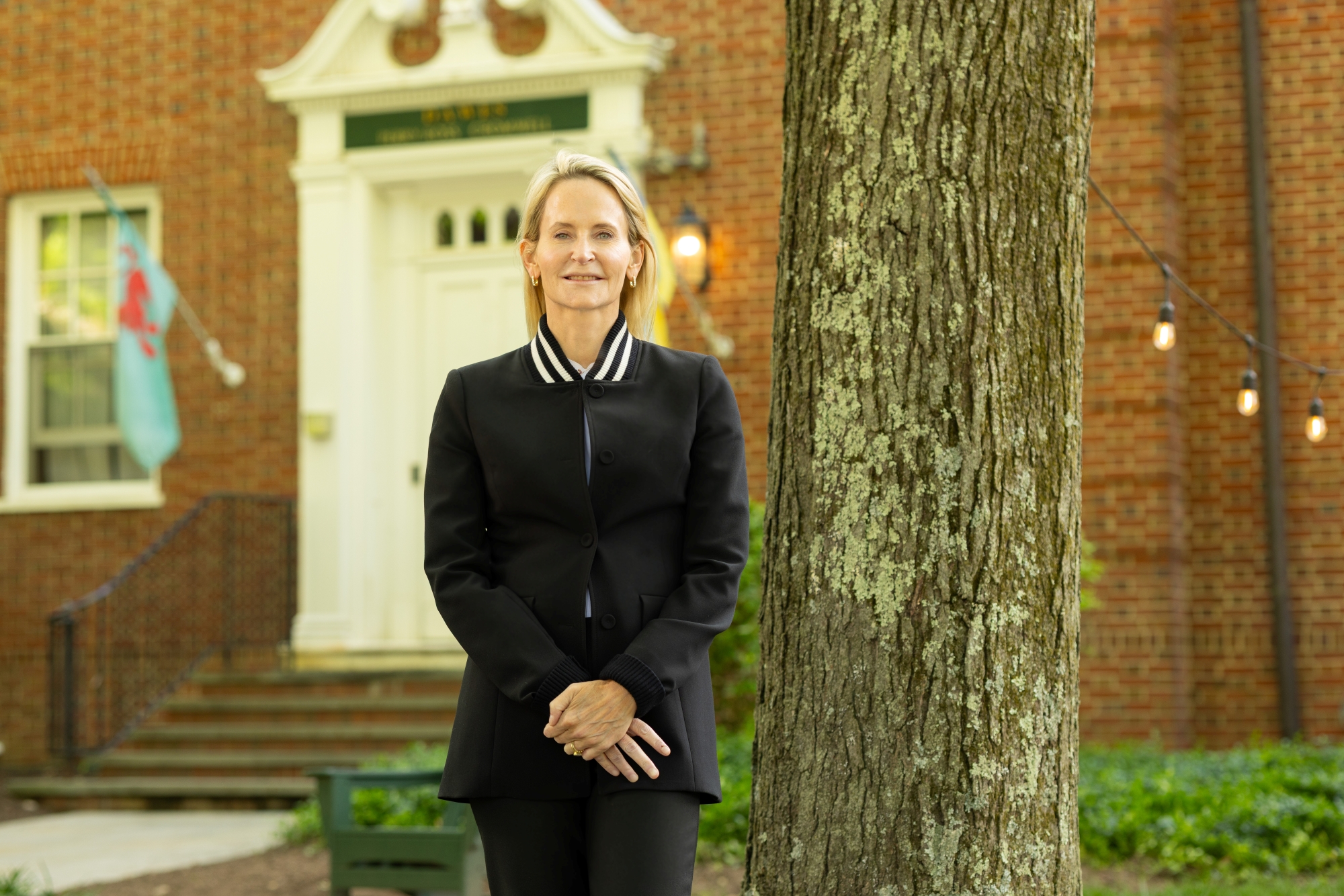 Celeste Mellet stands, smiling, with her hands clasped in front of her. She is wearing a black suit and a white shirt. She has shoulder-length blonde hair and a fair complexion. Behind her is a brick academic building.