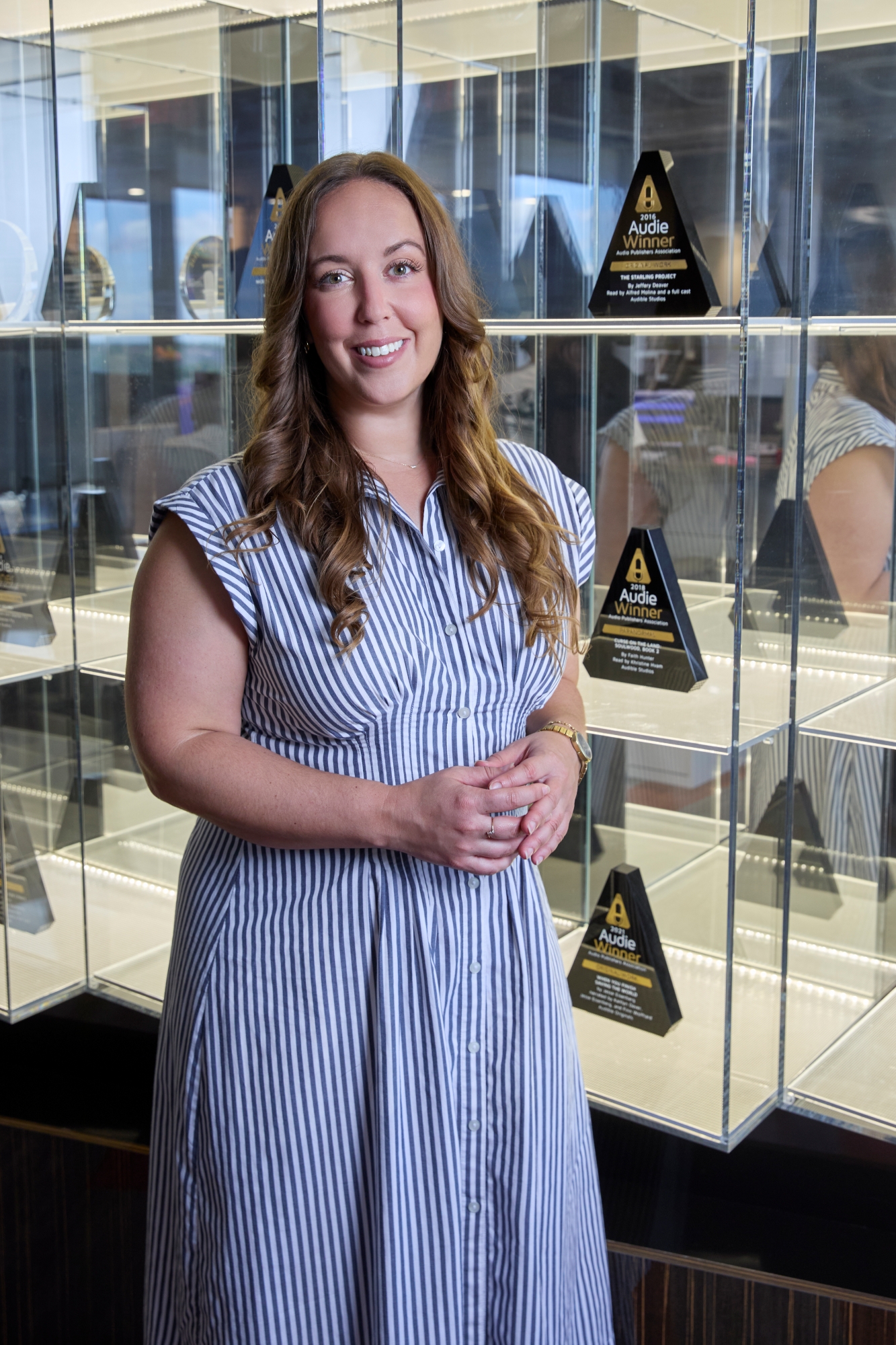 A woman with long brown hair stands before a glass trophy case with her hands clasped. The trophy case is filled with pyramid-shaped black trophies earned by her company, Audible books.