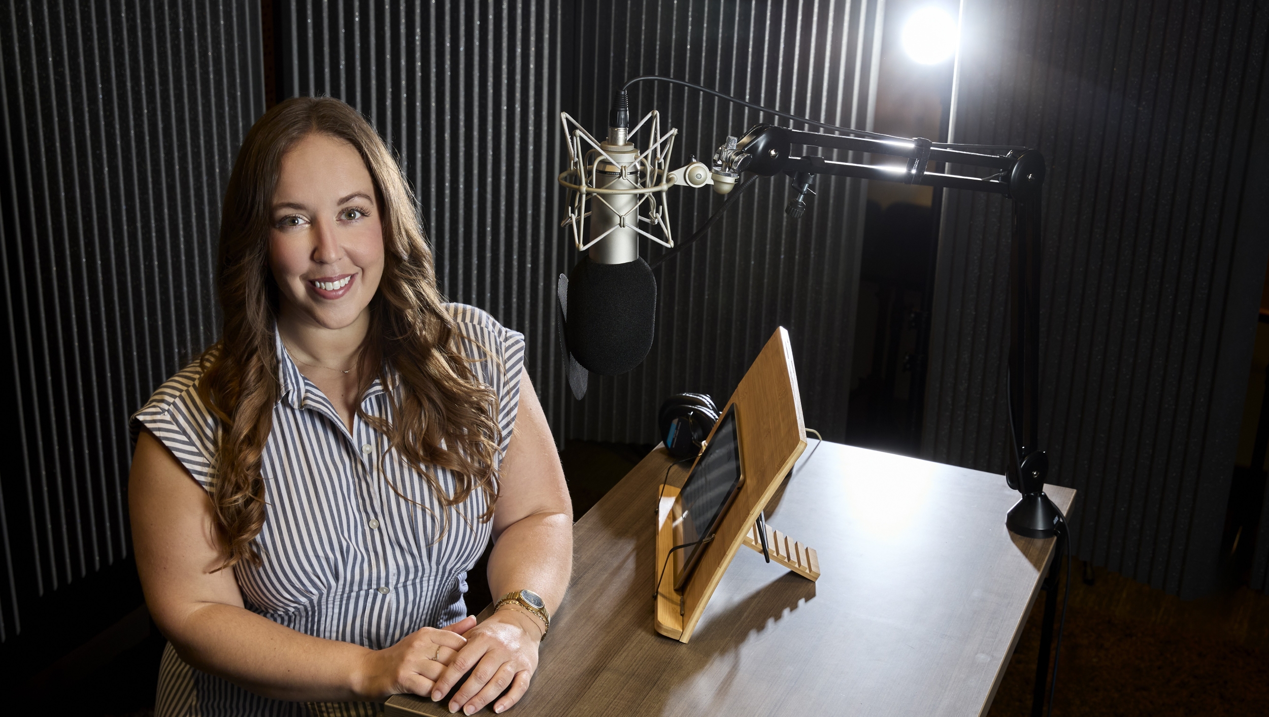 A woman with long brown hair stands before a hanging microphone in a recording studio. She is smiling with her hands placed on a brown wood-grain table.