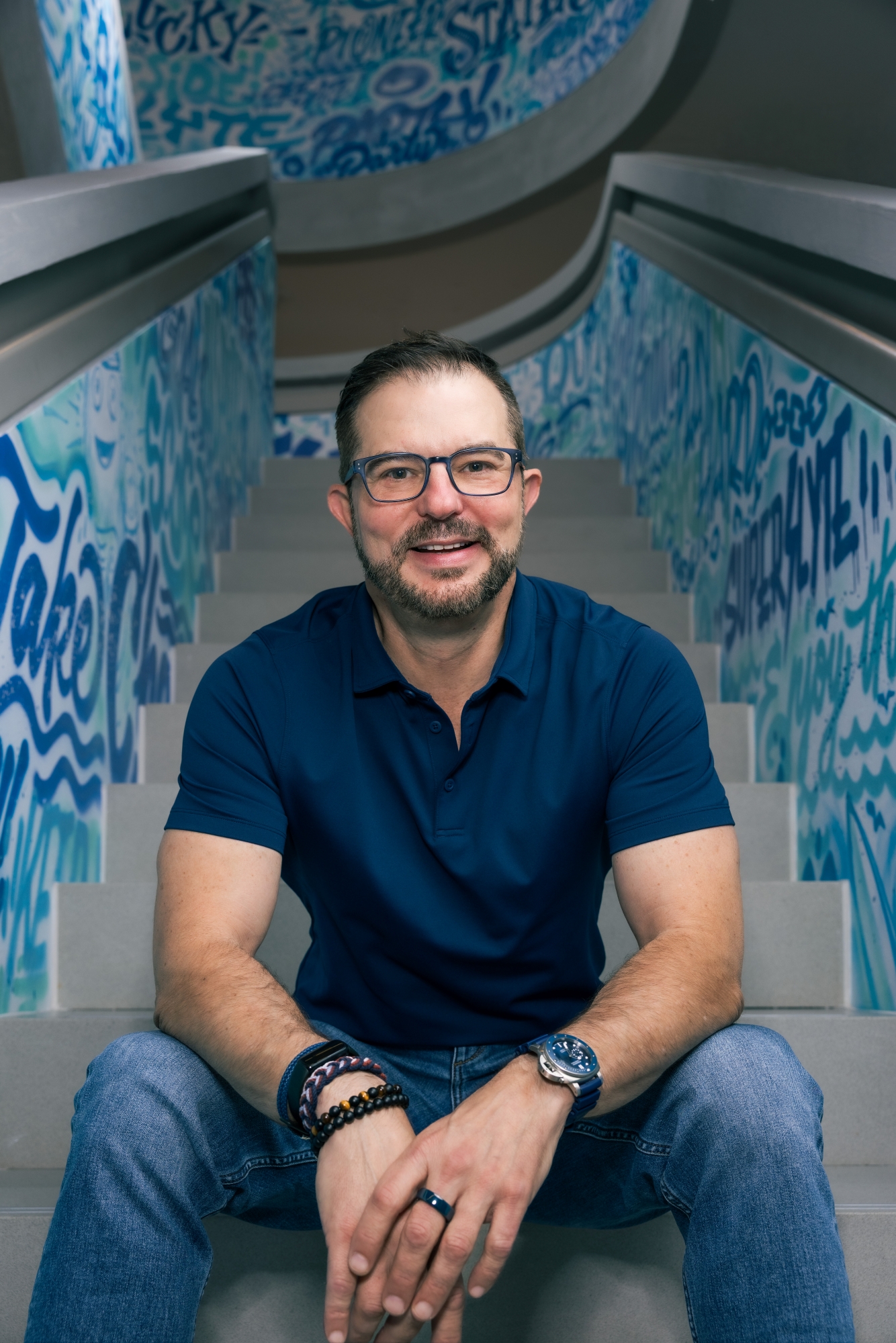 Clem Pappas, in a navy blue polo shirt and jeans, sits on the back of a blue couch with his hands resting on his thighs, sitting in a stairwell.