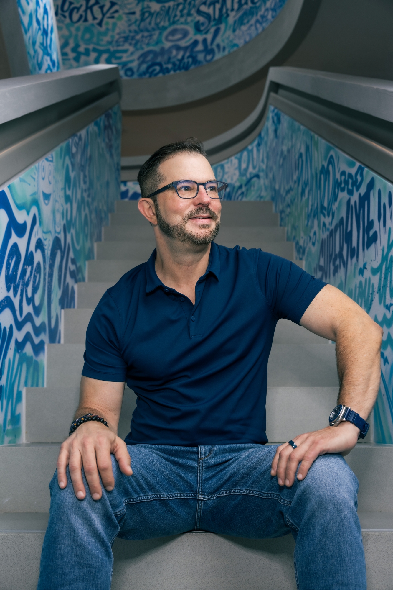 Clem Pappas, in a navy blue polo shirt and jeans, sits on the back of a blue couch with his hands resting on his thighs, sitting in a stairwell. He is looking up and to his left, as if into the distance.