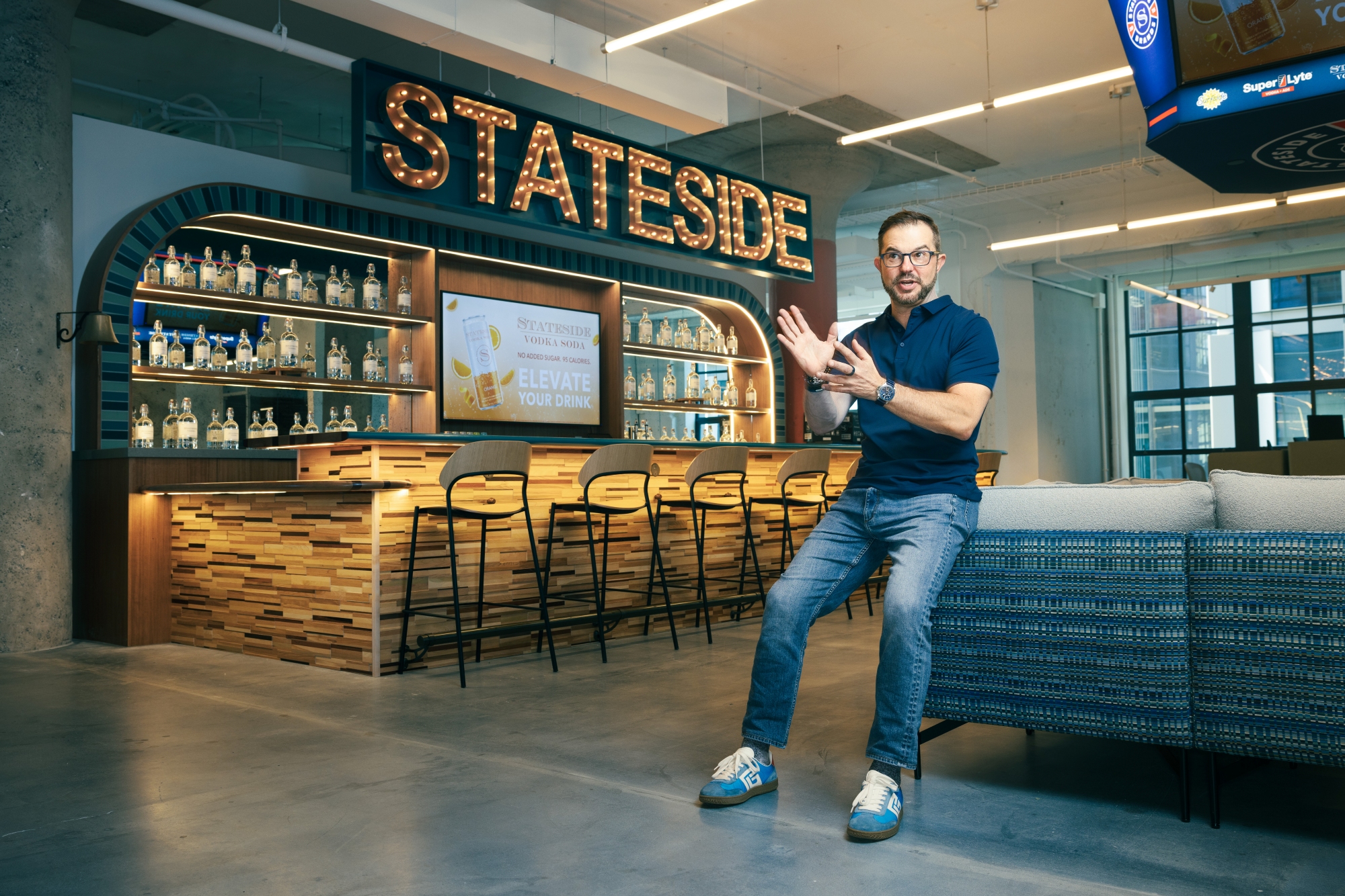 Clem Pappas, in a navy blue polo shirt and jeans, sits on the back of a blue couch, He is waving his hands as if to make point inside the bar area of his Stateside Brands headquarters. There is a long bar with a row of empty seats behind him, and in block letters formed by clear light bulbs, it spells out "STATESIDE" above the bartenders' area.