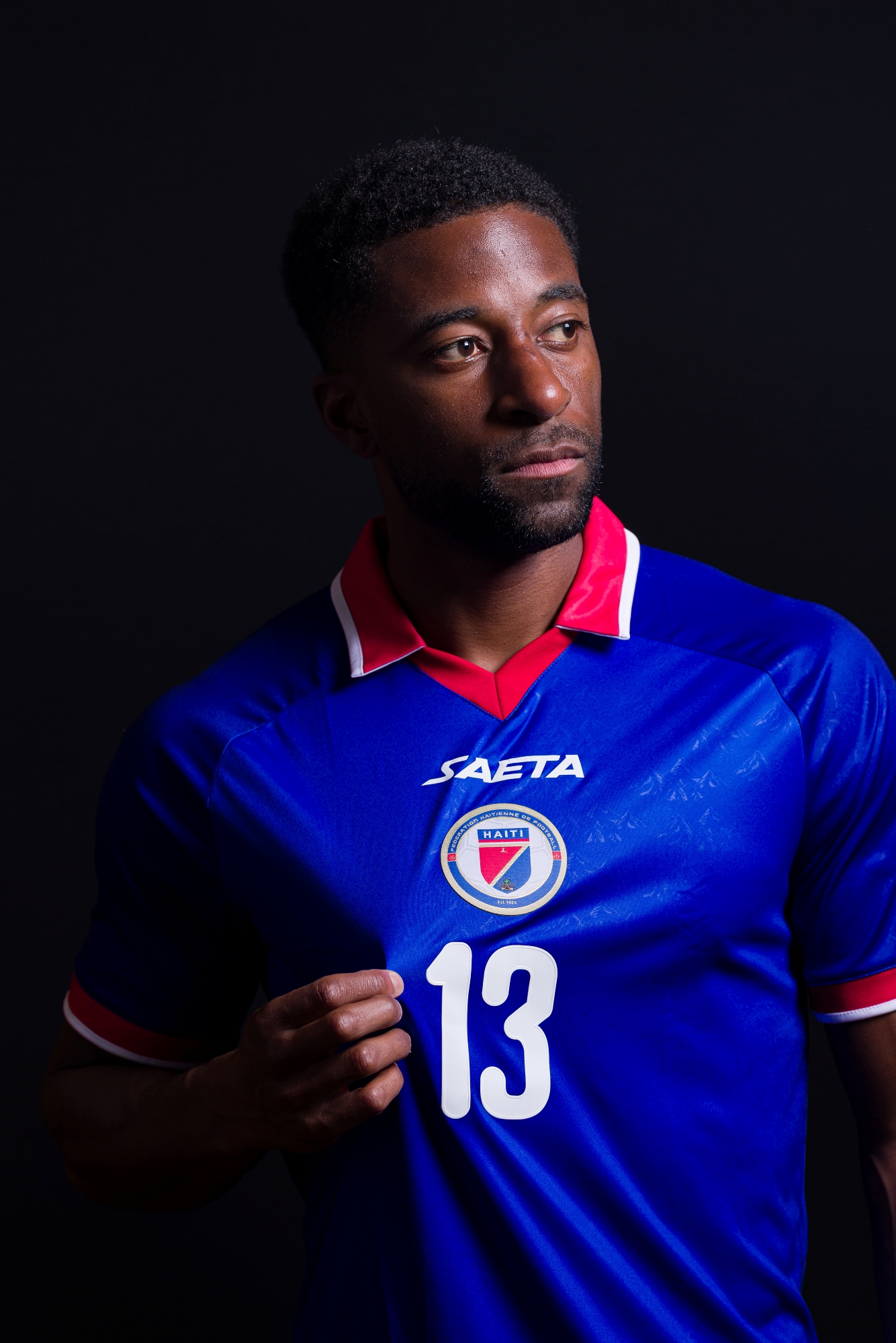 Duke Lacroix, wearing his royal blue Team Haiti jersey, pulls slightly at his number 13 in the center of his torso with his right hand. He is dramatically shaded on his right side as he looks seriously into the distance to his left side.