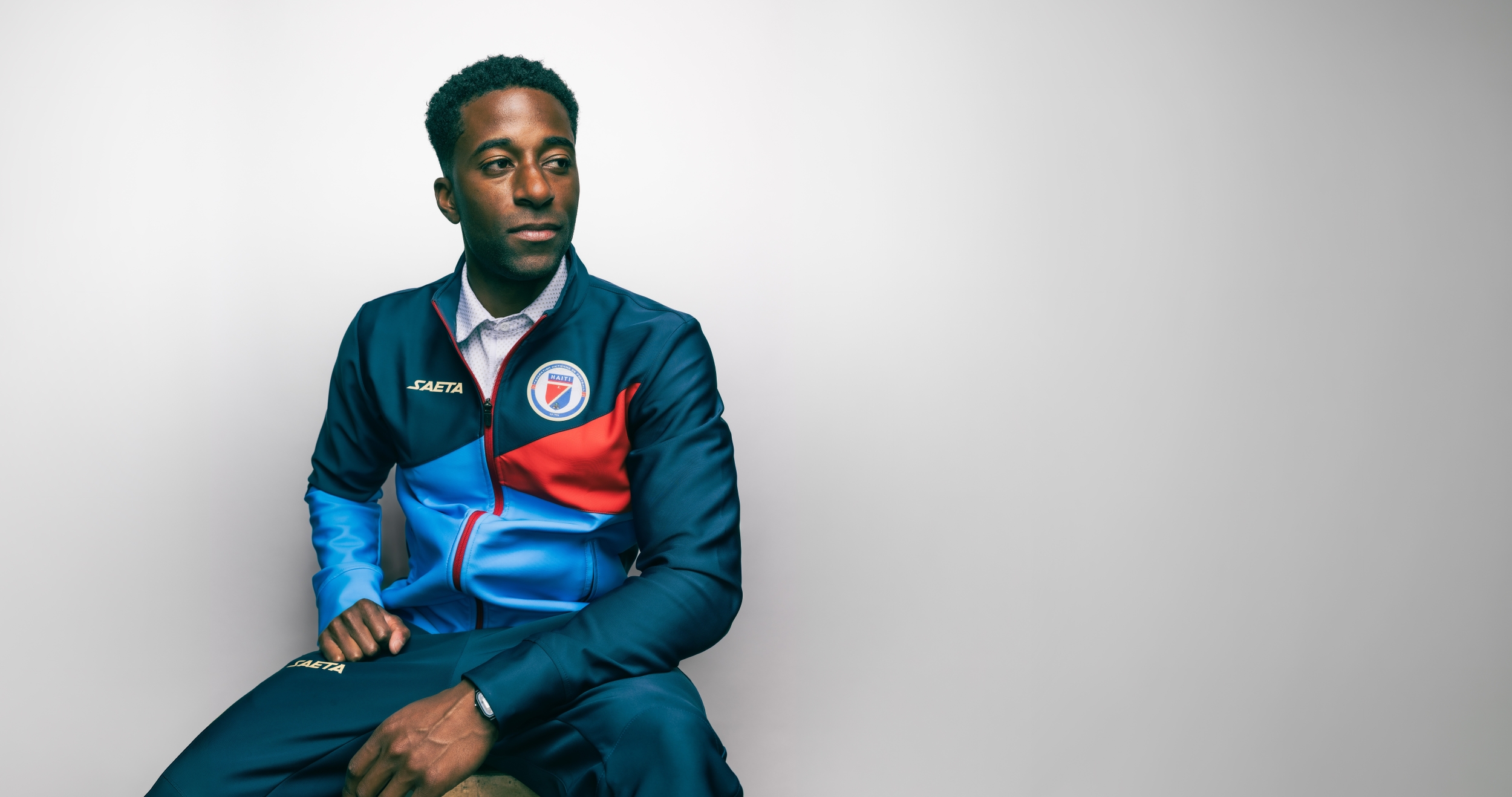 Duke Lacroix, wearing a dark blue, light blue, and red sweatsuit, sits on a stool with his hands resting in front of him. He is expressionless and looking slightly downward. The background is plain white.
