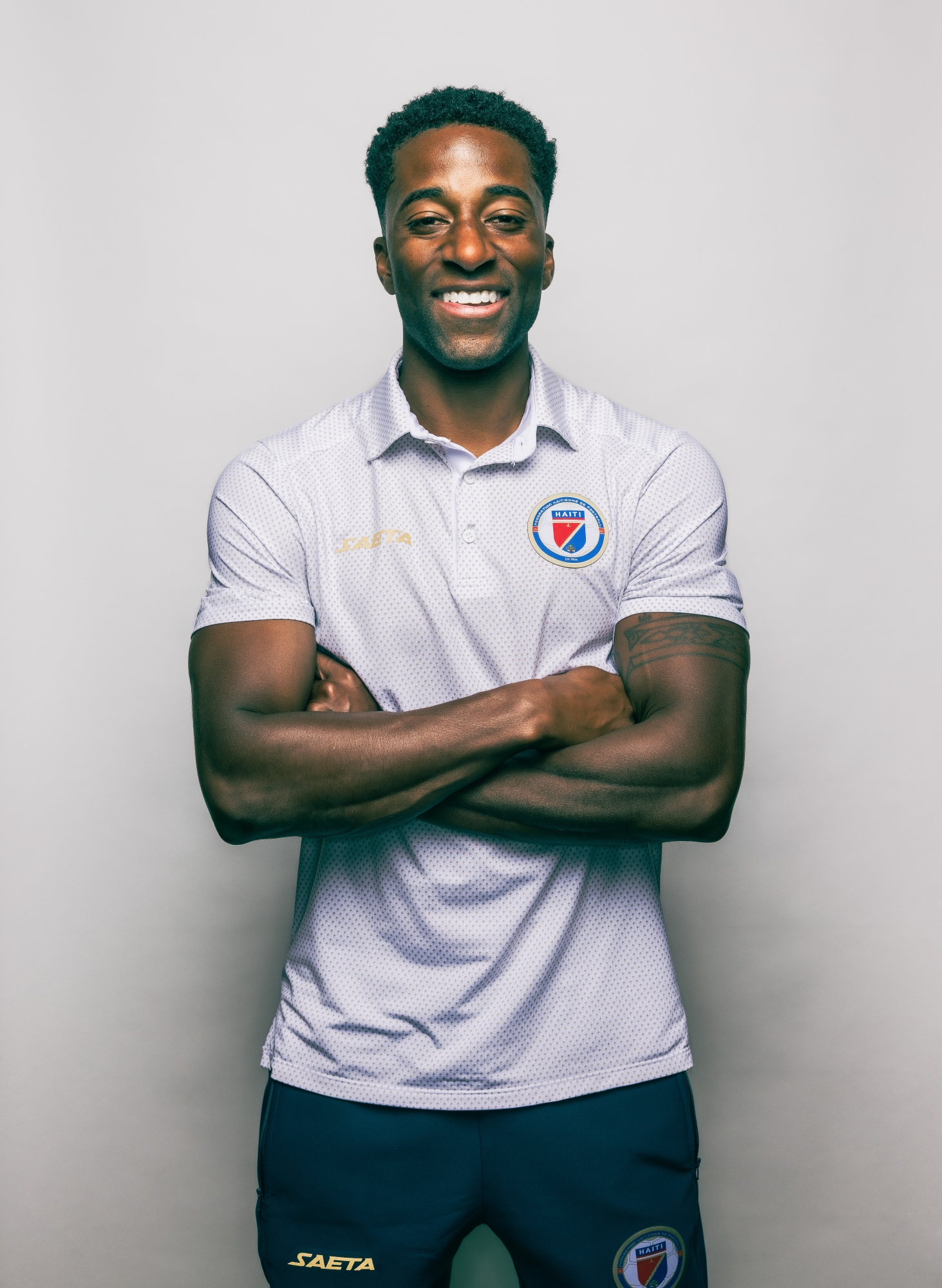 Duke Lacroix, wearing a white Team Haiti polo shirt, folds his arms and laughs while standing up against a plain white background.