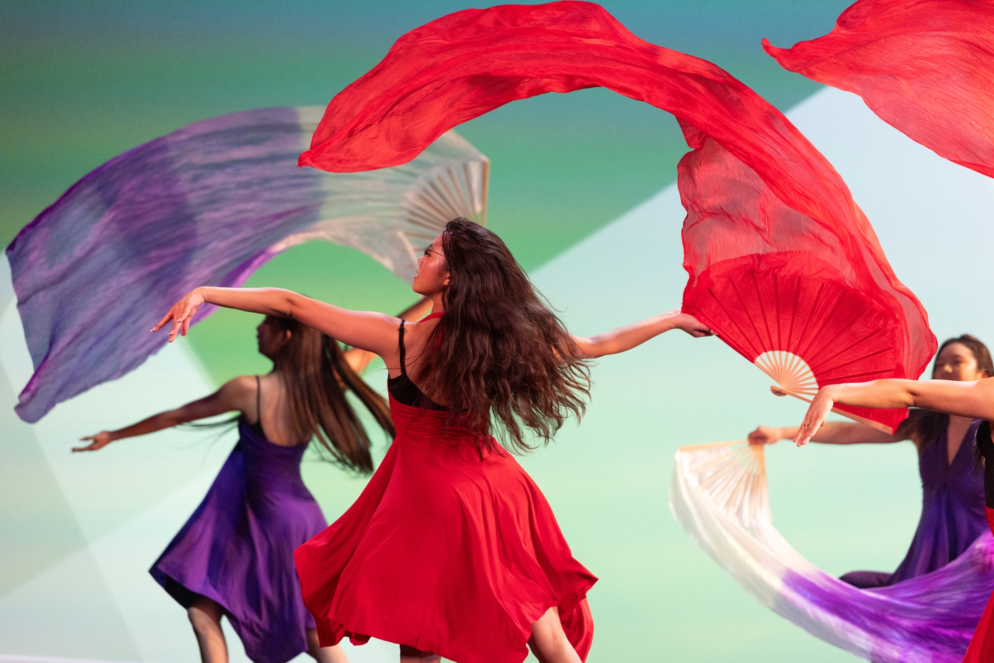 Girl dancers wearing dresses of different individual colors, dance as they wave flowing scarves of corresponding colors in unison.