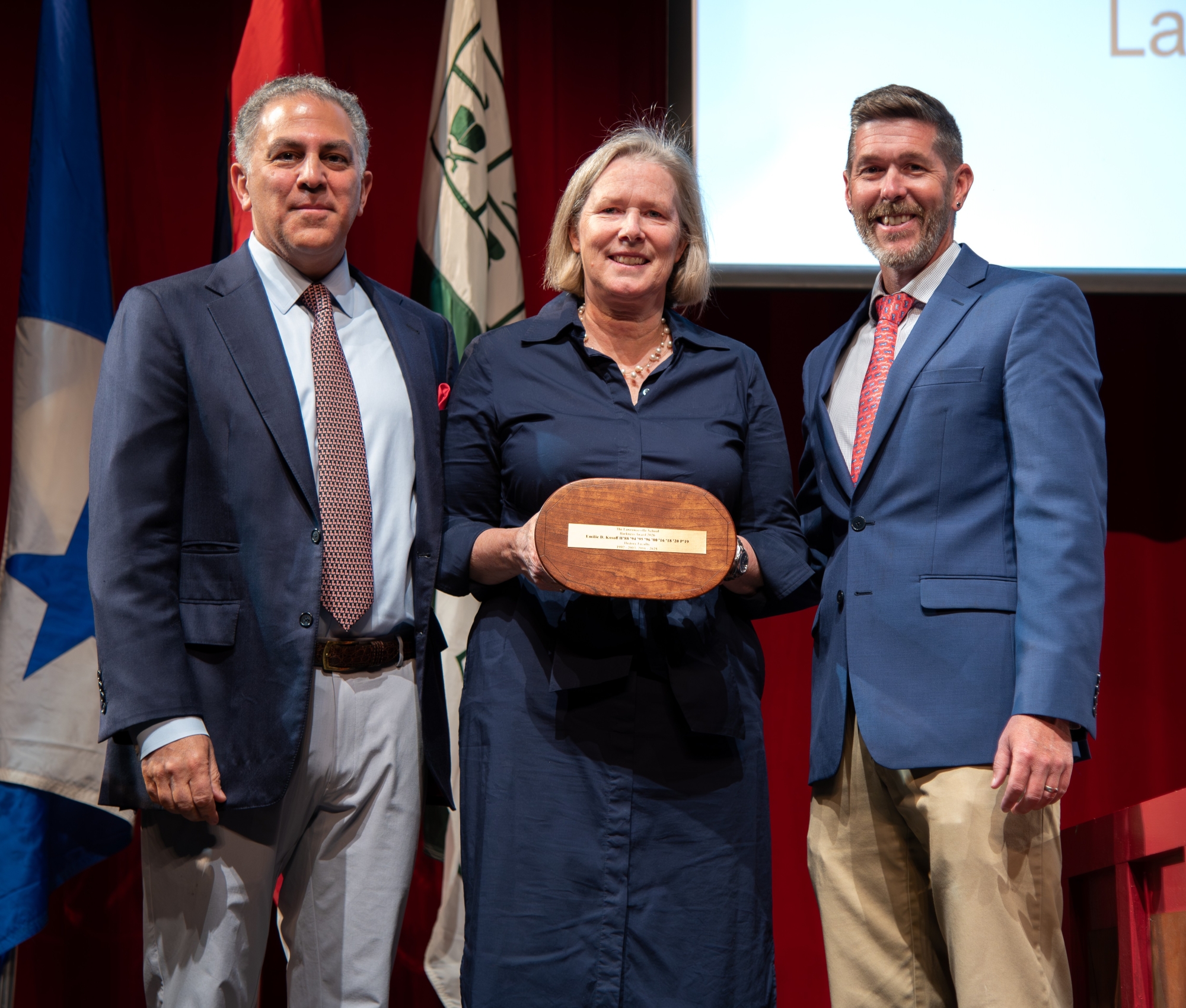 A woman in the middle holds a small replica of a Harkness table. She is flanked by two menwearing suits.