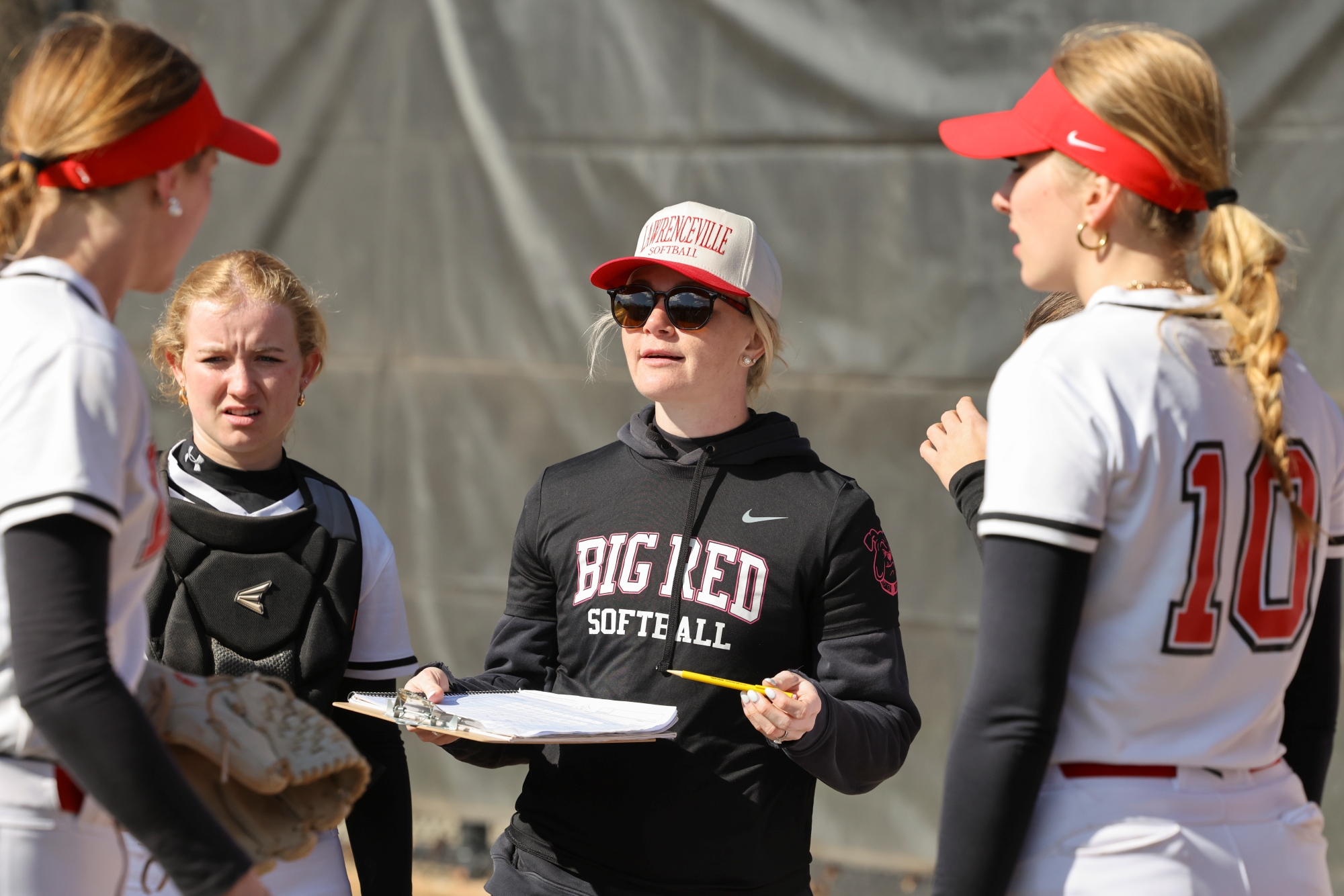 A woman coach dressed in black Lawrenceville softball gear and holding a clipboard addresses her players, who are wearing white softball uniforms.