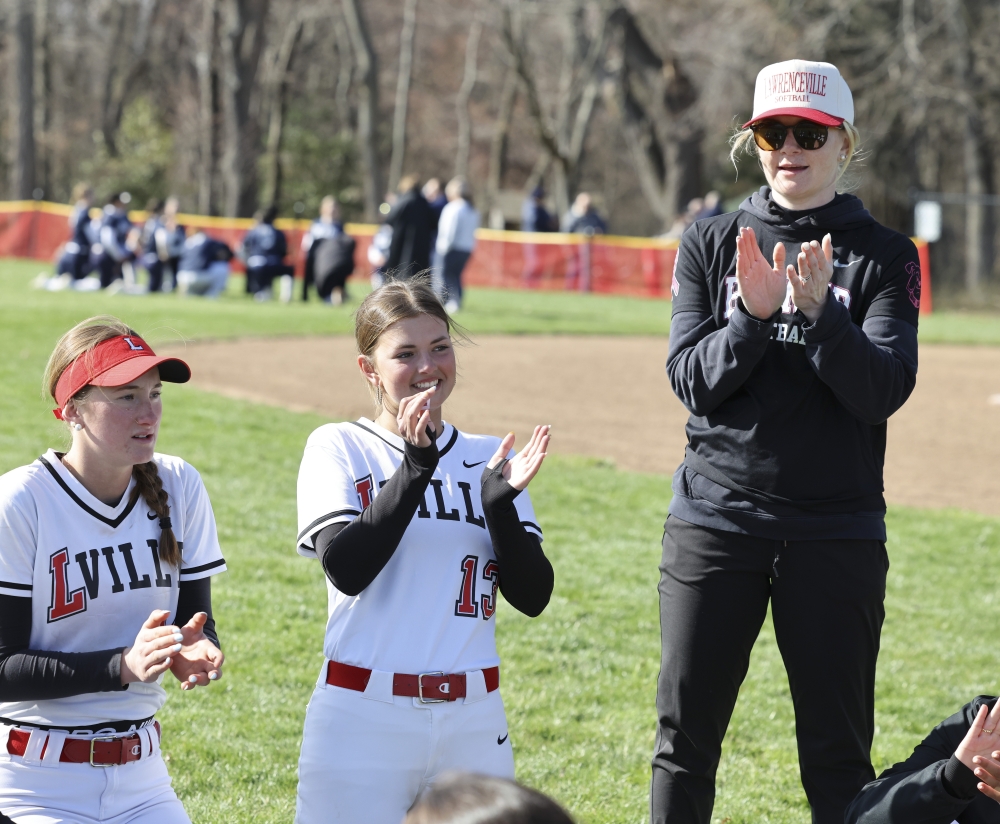 A woman coach dressed in black Lawrenceville softball gear applauds while her players, kneeling near her in white softball uniforms, applaud, too.