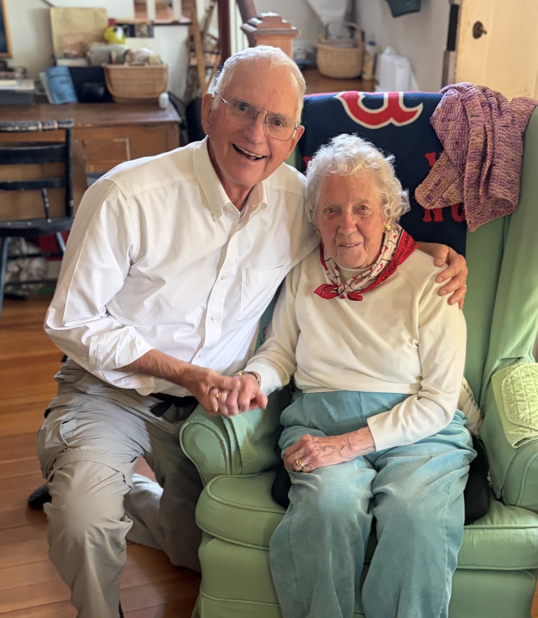 An older man with white hair, a white shirt and glasses kneels beside an older woman sitting in a green armchair. He has his left arm draped affectionately across her.