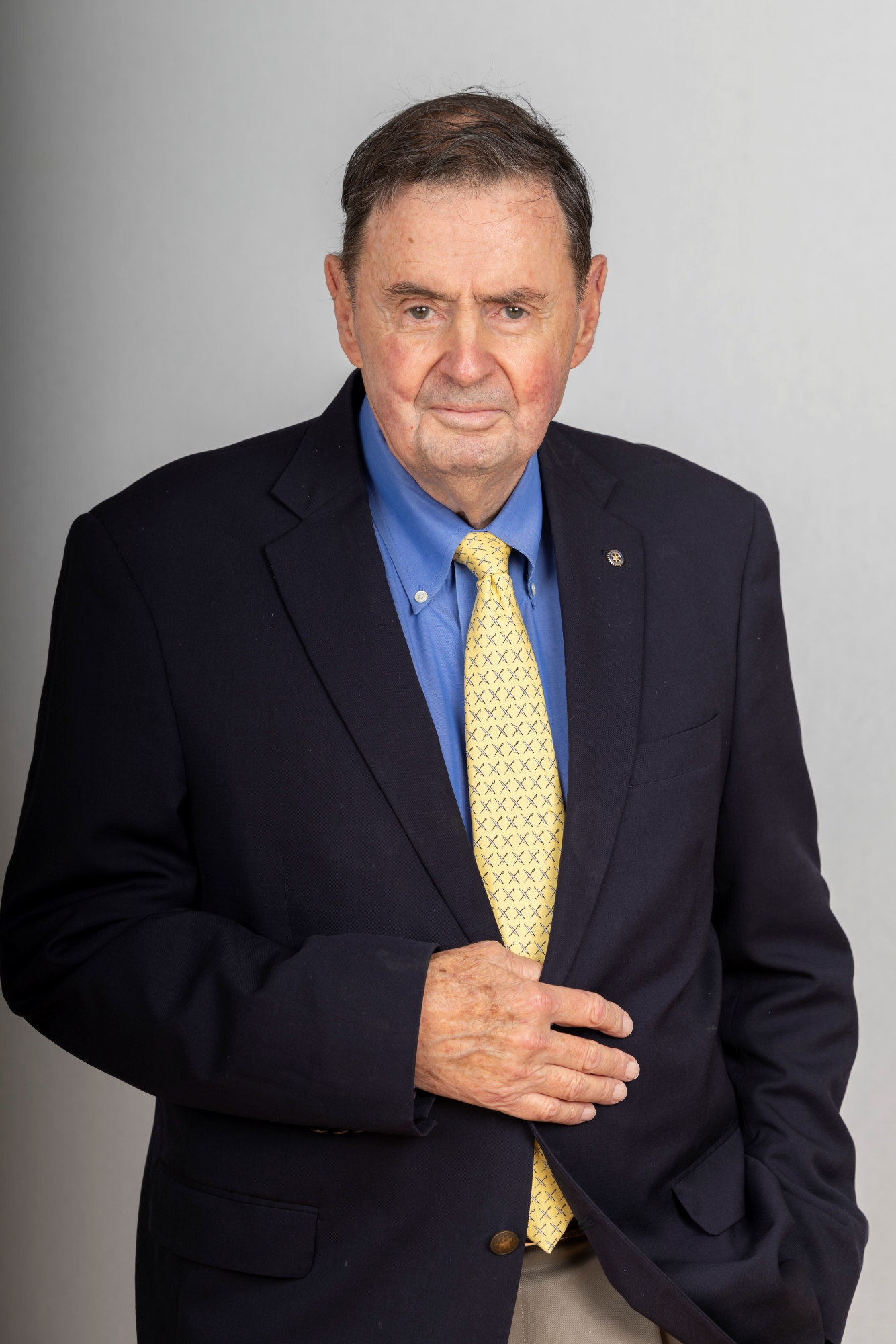 An older man with dark hair, a navy blue blazer, a French blue shirt and a yellow tie, poses for a portrait with his right hand across his torso.